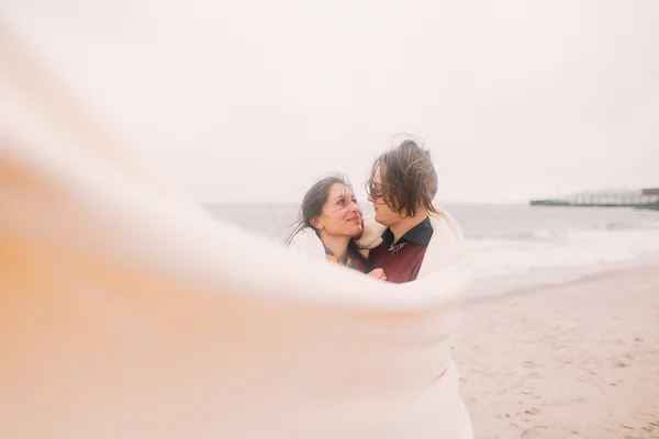 Krásný šťastný mladý pár se na sebe láskyplně podíval, zabalený do bílé přikrývky. Zimní moře na pozadí — Stock fotografie