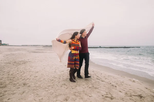 Attractive young happy couple wrapped in white blanket walking on the winter beach and looking at water. Vintage concept — Stock Photo, Image