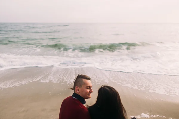 Beautiful couple sitting on beach, embracing and lovingly looking at each other. Winter seascape background. Tender moment. — Stock Photo, Image