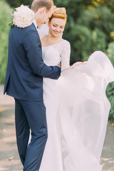 Boldog fiatal gyengéd vőlegény és a menyasszony pózol a parkban. Menyasszonyi esküvői csokor virág a kezében — Stock Fotó