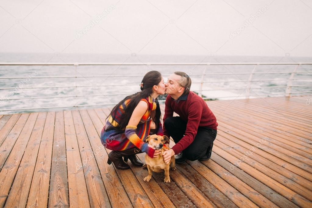 Young happy couple kissing each other and playing with dog on the rainy berth in autumn. Sea background