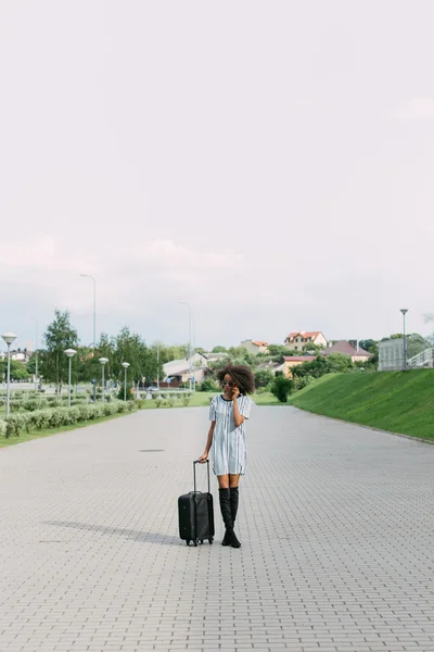 Moderna donna afro-americana in occhiali da sole che parla al cellulare e cammina con una valigia in mano . — Foto Stock