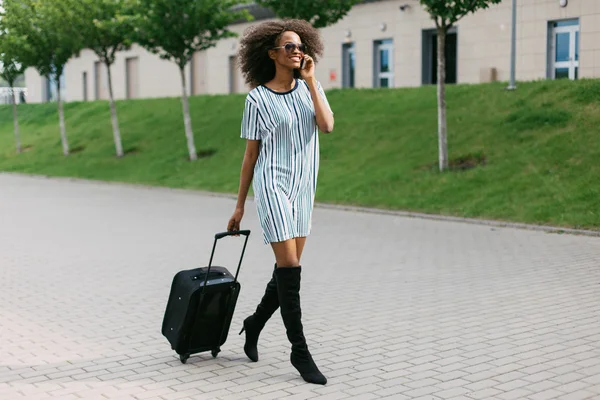 Vista laterale di una donna afro-americana in viaggio che cammina con valigia e telefono cellulare — Foto Stock