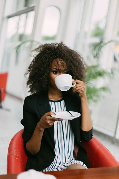 Приваблива афро-американська жінка сидить на столі в кафе. Вона тримає чашку кави і дивиться в напрямку — стокове фото