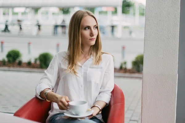 Krásná mladá blondýnka v bílé košili s kávovou šálkou, která se dívá na kavárnu — Stock fotografie