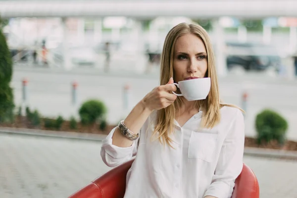 Close-up van mooie lachende jonge vrouw in shirt met blond haar en blauwe ogen drinken koffie of thee uit een witte beker — Stockfoto