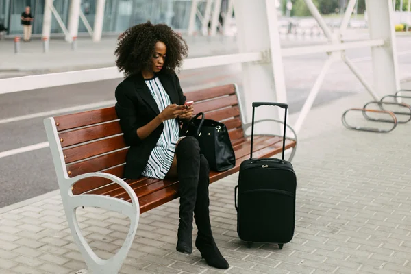 Bella donna afroamericana con valigia utilizzando smartphone mentre seduto sulla panchina vicino all'aeroporto . — Foto Stock