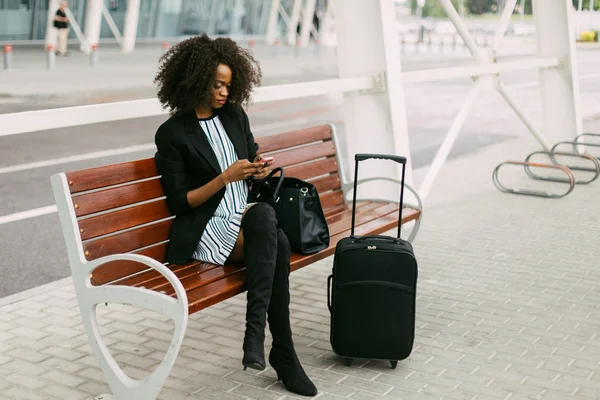 Giovane passeggera afroamericana con valigia nera che usa lo smartphone in attesa del suo volo. Concetto di viaggio aereo — Foto Stock