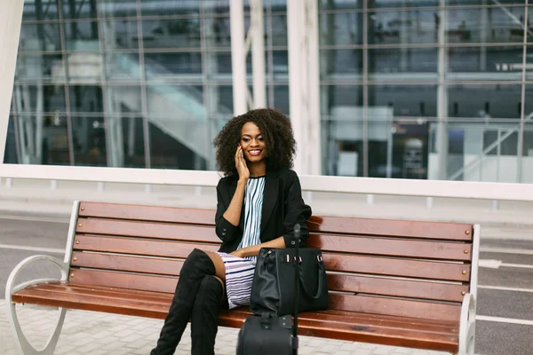 Retrato de uma mulher afro-americana sorridente esperando voo com bolsa e telefone celular — Fotografia de Stock