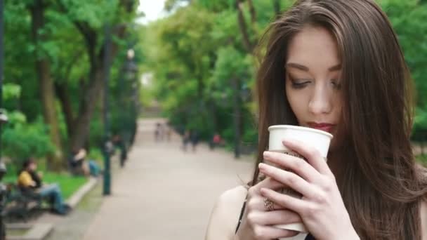 Hermosa chica morena con café para llevar caminando por la calle en el parque verde. Clima cálido y soleado. Al aire libre — Vídeos de Stock