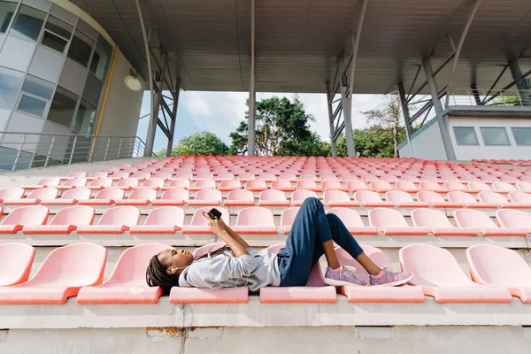 Bonito Africano americano jovem mulher ouvindo música com fones de ouvido deitado em um tribuno no estádio ao ar livre — Fotografia de Stock
