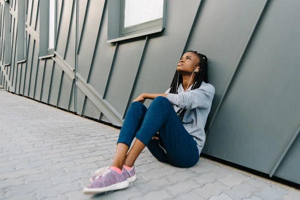 Sad young african american woman listening to melancholic music and looking up, sitting on the pavement — Stock Photo, Image