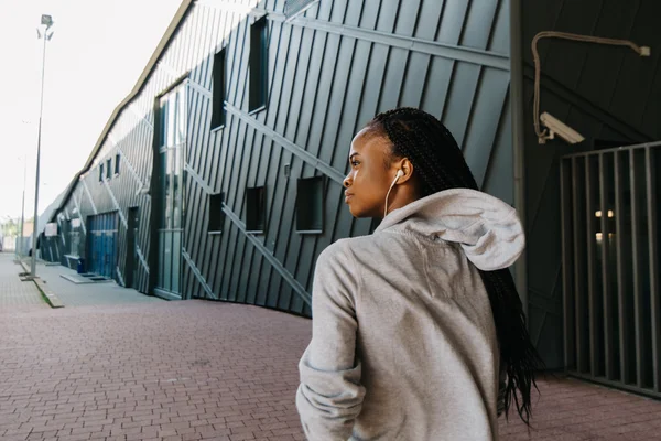 Retrato de una hermosa joven africana escuchando música con auriculares cerca de un edificio moderno, vista desde atrás — Foto de Stock