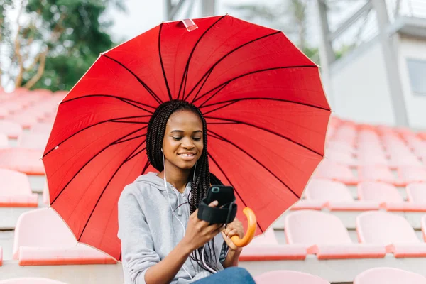 Smiling african american girl take a break and listen to music sitting on sport arena, holding red umbrella