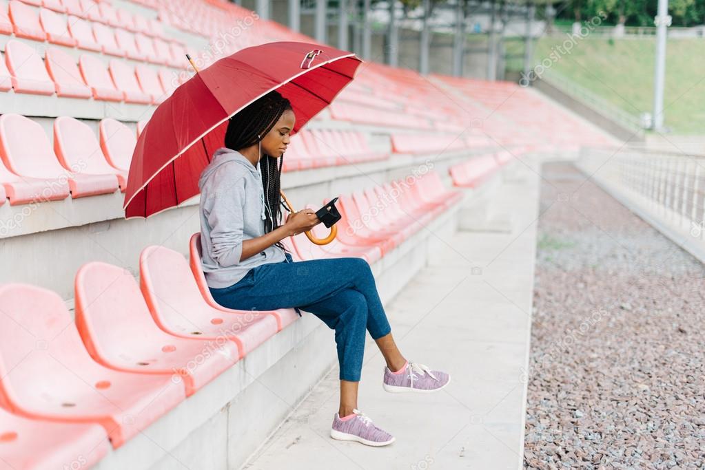 Full-length of single african american woman with red umbrella and mobile phone at the empty stadium