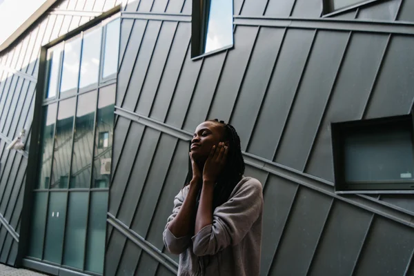 Gadis hitam muda di latar belakang kota mendengar musik, menutup telinga dengan tangannya — Stok Foto