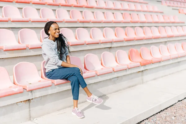 Full-length feliz menina afro-americana desportiva com fones de ouvido rindo na frente de bancos de estádio moderno — Fotografia de Stock