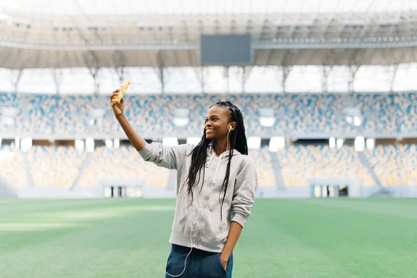 Esportes menina americana africana com fones de ouvido em pé na grama, mãos nos bolsos, faz selfie — Fotografia de Stock