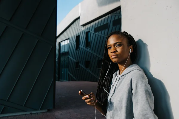 Donna afroamericana che ascolta musica in cuffia su edifici moderni sfondo, primo piano — Foto Stock