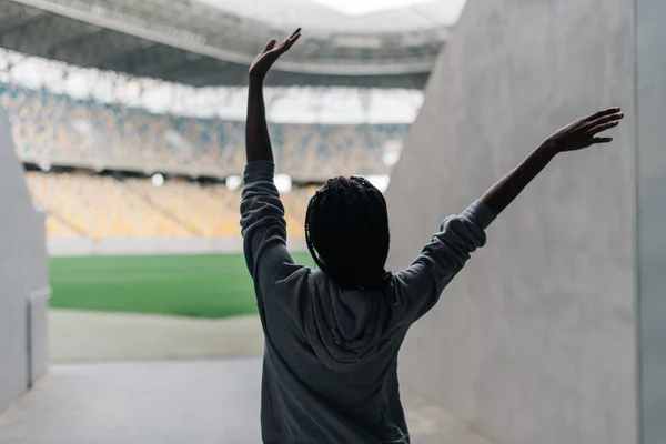 Gelukkig Afro-Amerikaanse vrouw staande onder lege stadion vieren, haar armen verhoogd ponsen de lucht met handen, uitzicht vanaf rug — Stockfoto