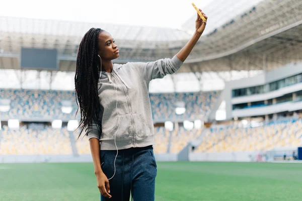 Joven hermosa afroamericana deportista haciendo foto selfie en el teléfono móvil después de entrenar al aire libre y sonriendo. Fit mujer es en grande agradable estadio moderno — Foto de Stock