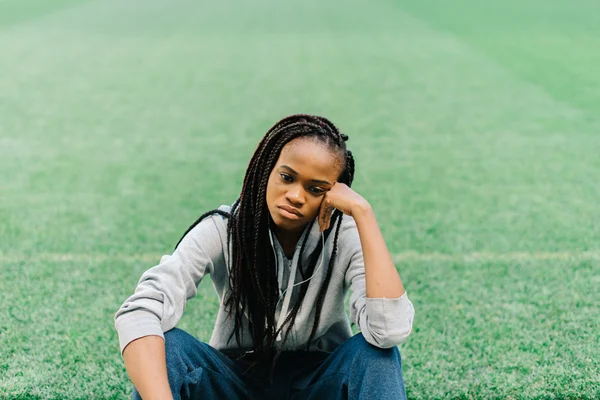 Mulher americana africana esportiva sentada com a mão na cabeça e sentindo-se desapontada contra a grama verde no estádio — Fotografia de Stock