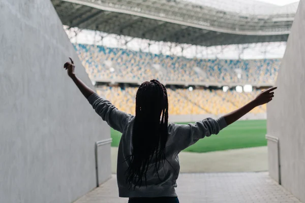 Happy african american woman standing among empty stadium celebrating, her arms raised punching the air with hands, view from back