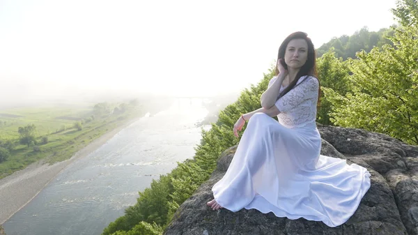 Portrét brunetka mladá nevěsta v bílé svatební šaty, sedícího u řeky a těší se v okamžiku — Stock fotografie