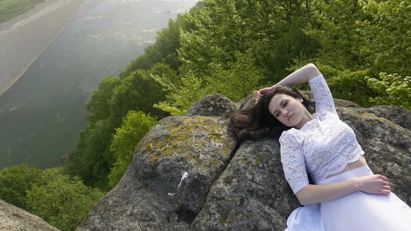 Retrato da jovem noiva pensativa em um vestido de noiva branco deitado sobre as rochas sozinho perto do rio — Fotografia de Stock