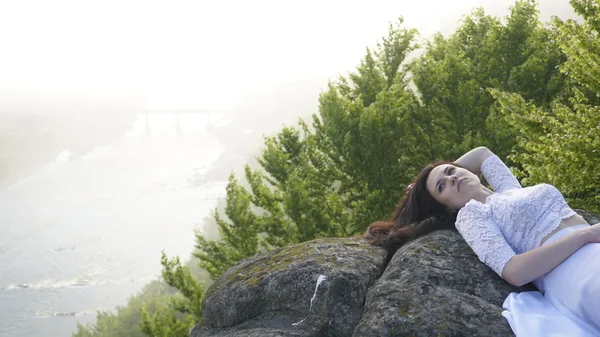 Retrato de jovem noiva pensativa deitada nas rochas sozinha e olha para o outro lado. Rio em nevoeiro no fundo — Fotografia de Stock