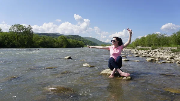 Portret van lachende mooie jonge vrouw zittend op de steen en tonen vredesteken. Karpaten berg rivier achtergrond — Stockfoto