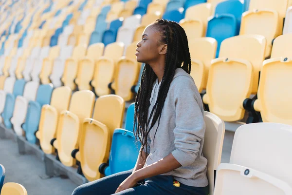 Emocionada mujer afroamericana en ropa deportiva gris mirando hacia otro lado sentada en gradas esperando un espectáculo, celebración —  Fotos de Stock
