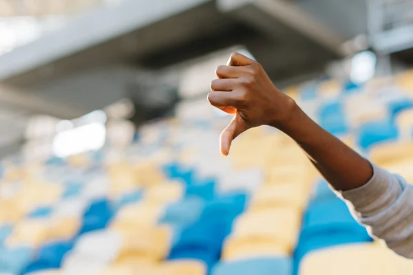 Gesto, conceito de partes do corpo - mãos de mulher africana mostrando polegares para baixo. Assentos de estádio de plástico azul e amarelo como fundo — Fotografia de Stock