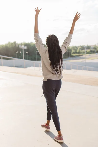 Vista lateral trasera de la mujer feliz con auriculares y manos levantadas caminando al aire libre en tiempo soleado —  Fotos de Stock