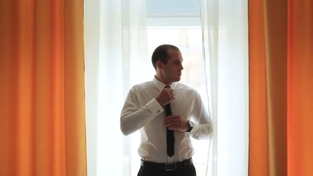 Чоловік у білій сорочці зав'язує краватку перед вікном з помаранчевими шторами — стокове відео