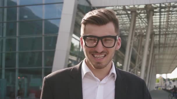 Close-up face de confiante bonito sorridente empresário ajustando óculos e olhando para a câmera. Contexto do aeroporto ao ar livre, conceito de negócio — Vídeo de Stock