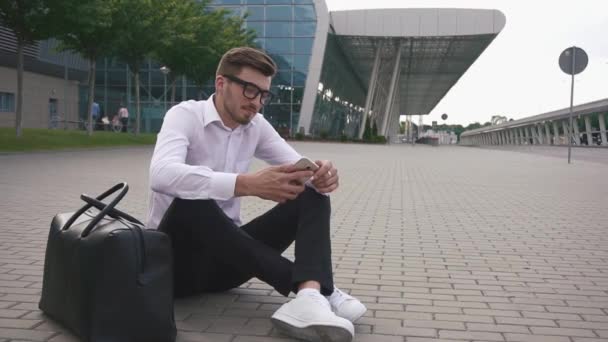 Hombre de negocios barbudo guapo y seguro en gafas en el aeropuerto sentado en el suelo con equipaje y teléfono y esperando su vuelo — Vídeo de stock