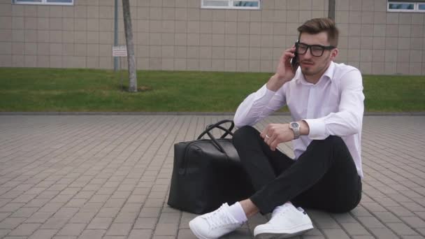 Bonito jovem empresário de óculos falando no telefone ao ar livre enquanto sentado no chão na rua — Vídeo de Stock