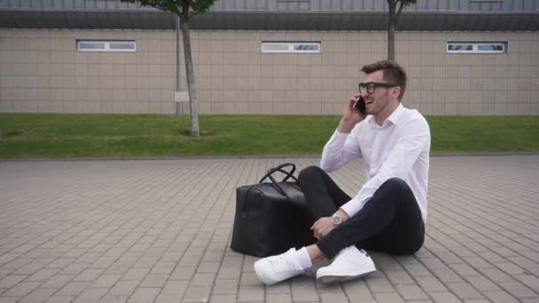 Eleganter junger Geschäftsmann mit Brille, der im Freien lacht und telefoniert, während er auf dem Boden an der städtischen Straße sitzt — Stockvideo