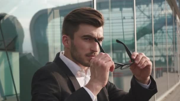 Primer plano retrato de un atractivo hombre de negocios toma gafas y estar preocupado y reflexivo mientras está de pie junto a los edificios de oficinas en una plaza clásica de la ciudad . — Vídeos de Stock