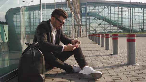 Bello uomo d'affari barbuto nella stazione aeroportuale digitando un messaggio di testo sul suo telefono cellulare e seduto a terra accanto all'ingresso — Video Stock