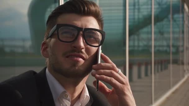 Крупним планом красивий бородатий бізнесмен розмовляє по смартфону, сидячи в аеропорту і чекаючи на політ. Скляний фон стіни — стокове відео