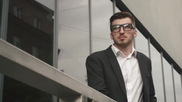 Joven hombre de negocios guapo y seguro esperando su vuelo al aire libre en el aeropuerto. Modelo de hombre de negocios masculino en gafas en el fondo de pared de vidrio del aeropuerto urbano — Vídeos de Stock