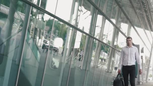 Joven empresario confiado en gafas y con maleta en el moderno fondo de pared de cristal terminal del aeropuerto. Concepto de chico viajero — Vídeo de stock