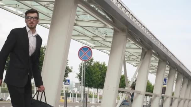 Joven hombre de negocios en gafas y con barba corriendo a su avión con gran maleta en cámara lenta. Fondo urbano de la ciudad — Vídeos de Stock