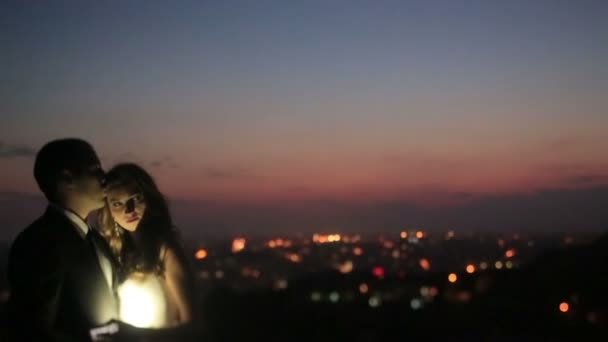 Charmant couple de mariage sur la colline du coucher du soleil sur les lumières de la ville du soir, illuminé par une lampe de poche à la main des mariées — Video