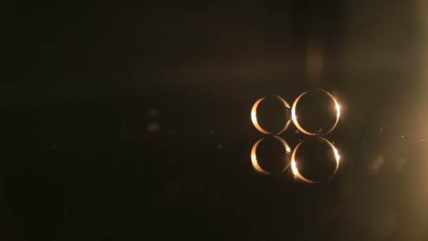 Two golden wedding rings on the mirroring surface, with dark background illuminated with warm light — Stock Video