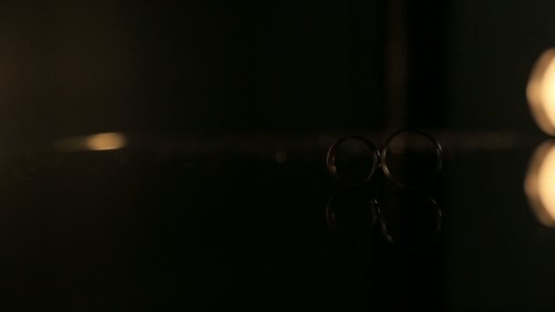 Dos anillos de boda dorados en la superficie del espejo, con fondo oscuro iluminado por una lámpara móvil con luz cálida — Vídeo de stock