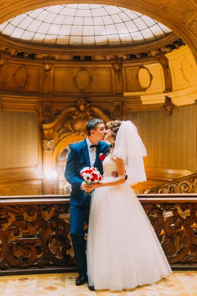 Enloved noivo beijando sua noiva encantadora perto de escadas de madeira velha com o fundo do interior de luxo — Fotografia de Stock