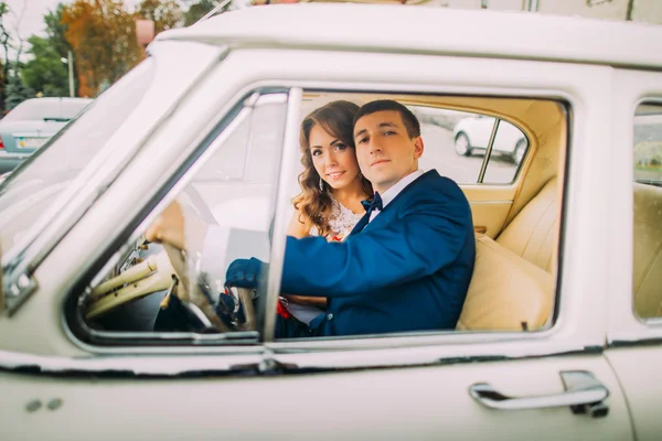 Feliz casal recém-casado sentado em carro antigo. As mãos dos noivos estão no volante — Fotografia de Stock